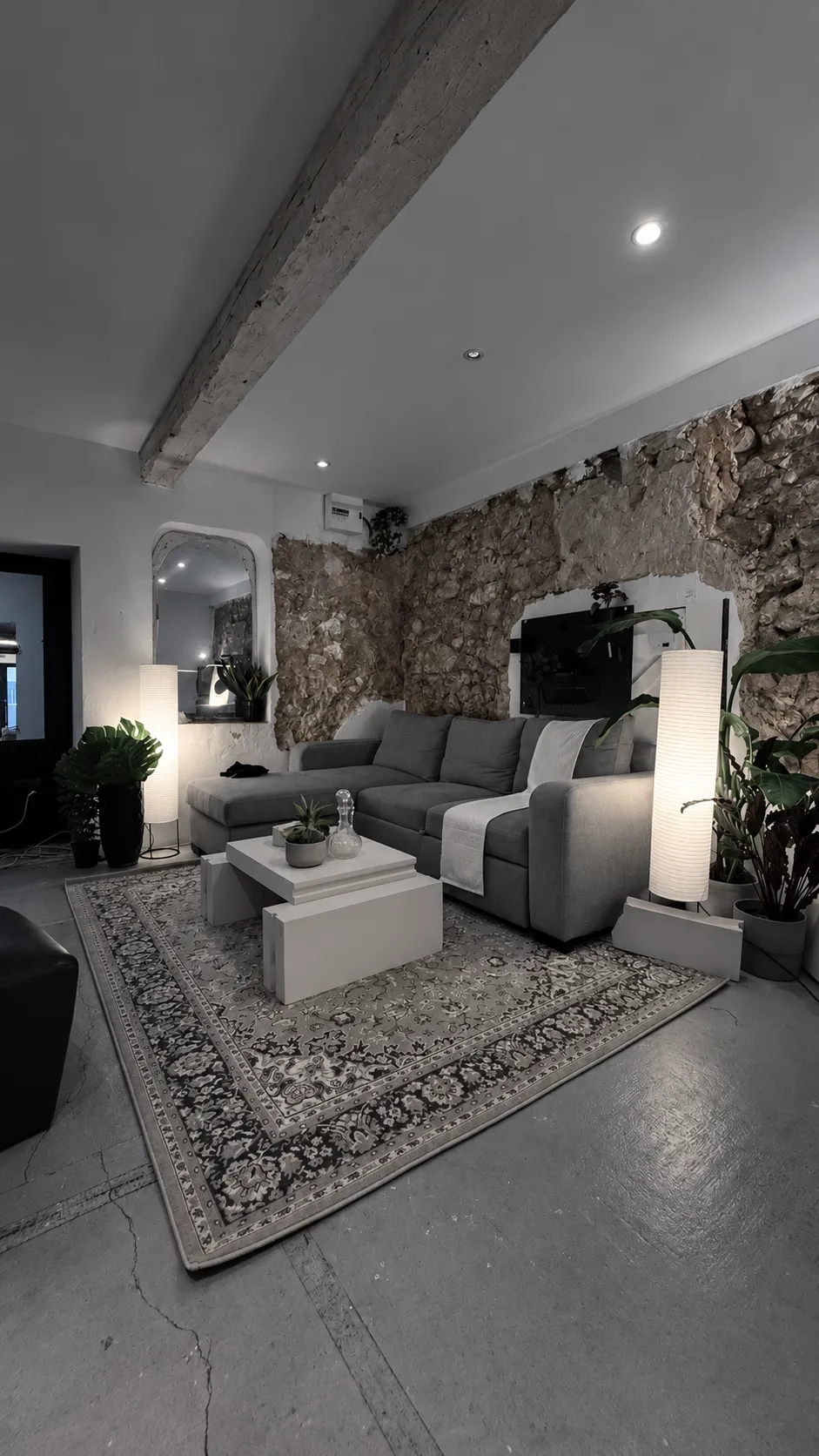 Le coin salon : canapé gris, tapis, lampes en papier et plantes devant un mur en pierre